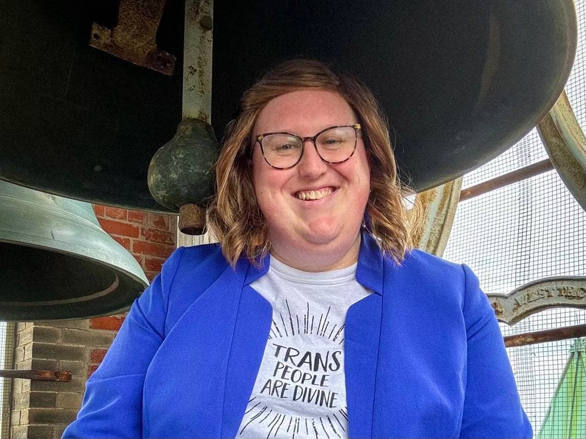 Katelyn MacDonald wearing a blue blazer with a t-shirt that reads "Trans People Are Divine" while standing underneath a large church bell