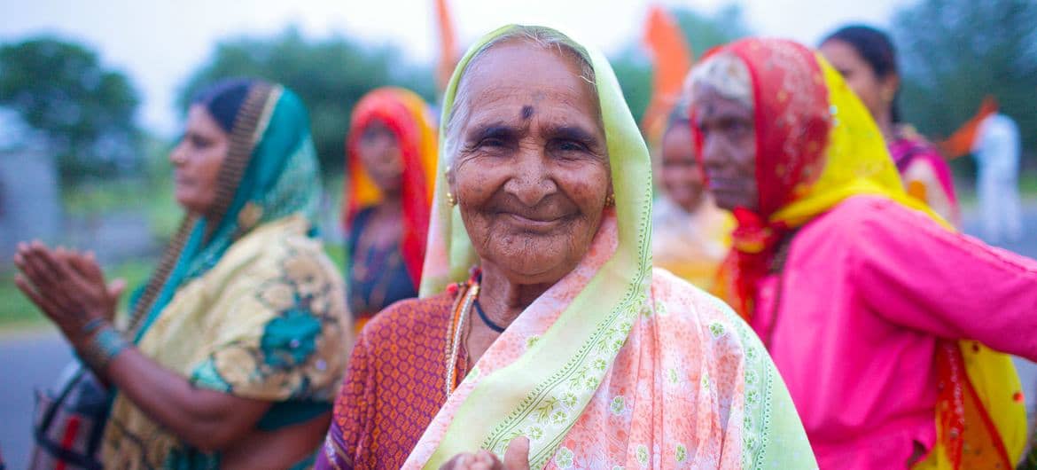 An elderly woman wearing a light green and pink headscarf and patterned clothing smiles at the camera. Several other women in colorful headscarves and garments stand behind her outdoors.