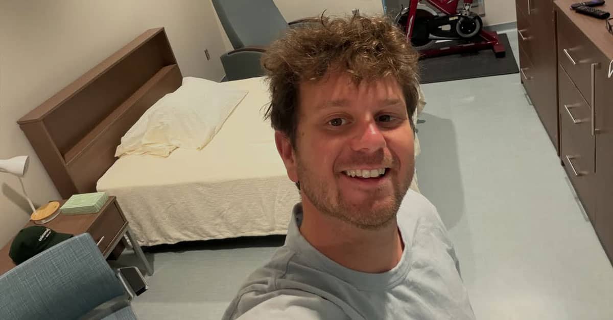 A person with dark blonde hair smiling, taking a selfie with a hotel-looking room behind him