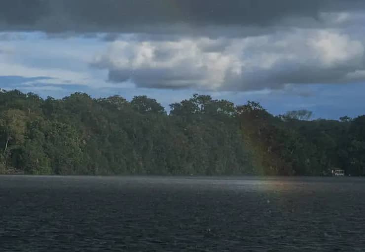 A dark body of water with dense green forest lining its shore under a partly cloudy sky with gray and white clouds, a subtle rainbow is visible over the water in front of the treeline.