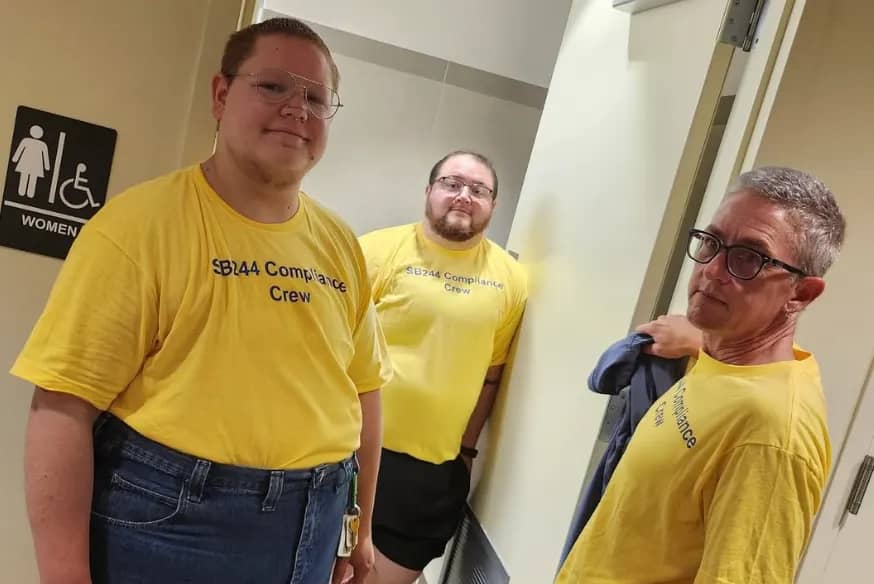 Three people wearing yellow "SB144 Compliance Crew" t-shirts stand in a hallway near a women's restroom sign. 