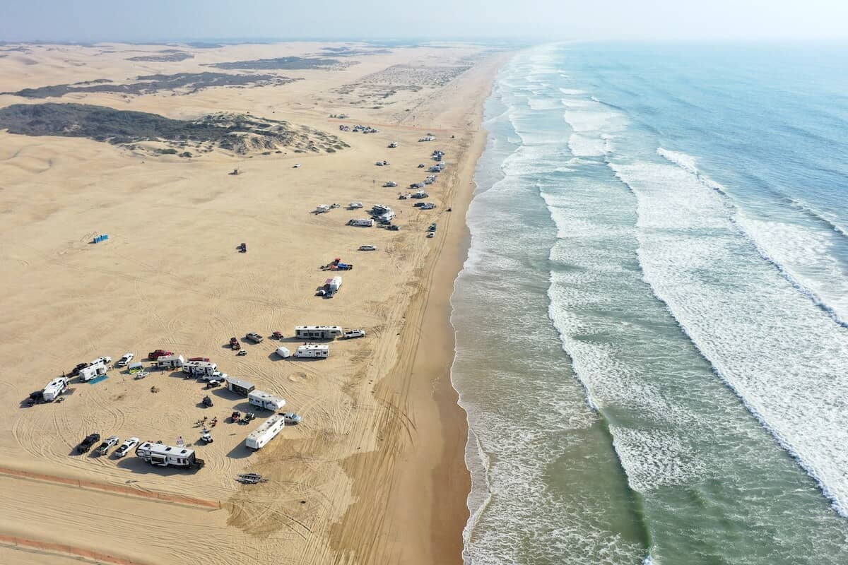 Aerial photo of waves crashing onto a tan sandy beach with campers, cars, and people on the coastline
