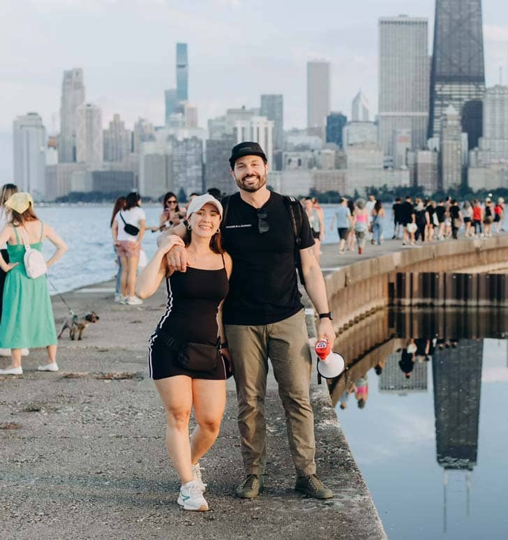 Two people standing near a body of water with a city scape in the background