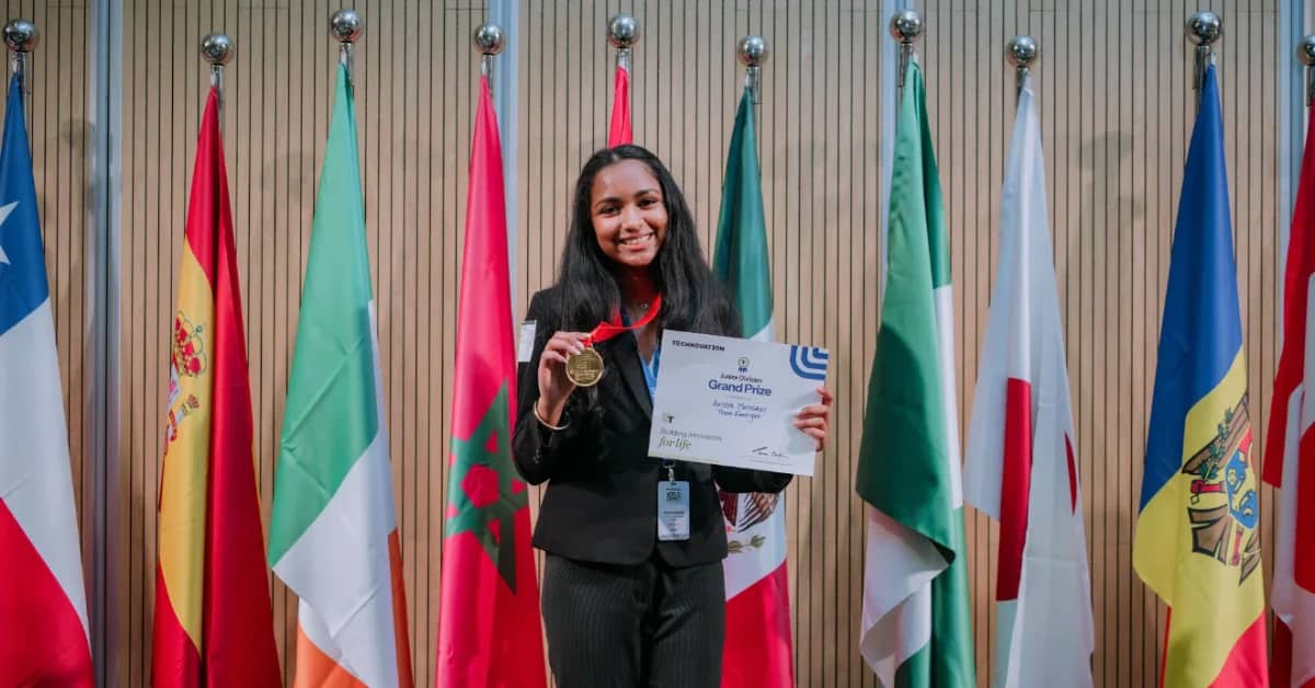 A woman in a black blazer holds up a gold medal and a certificate reading "Grand Prize" in front of a backdrop of international flags.