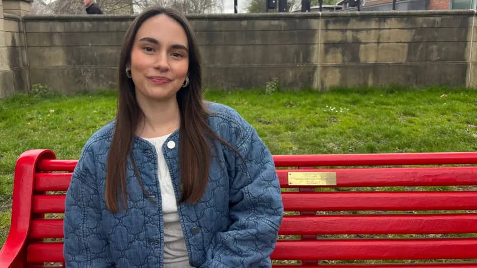 Young woman with brown hair wearing a white shirt and denim jacket sitting on a bright red bench