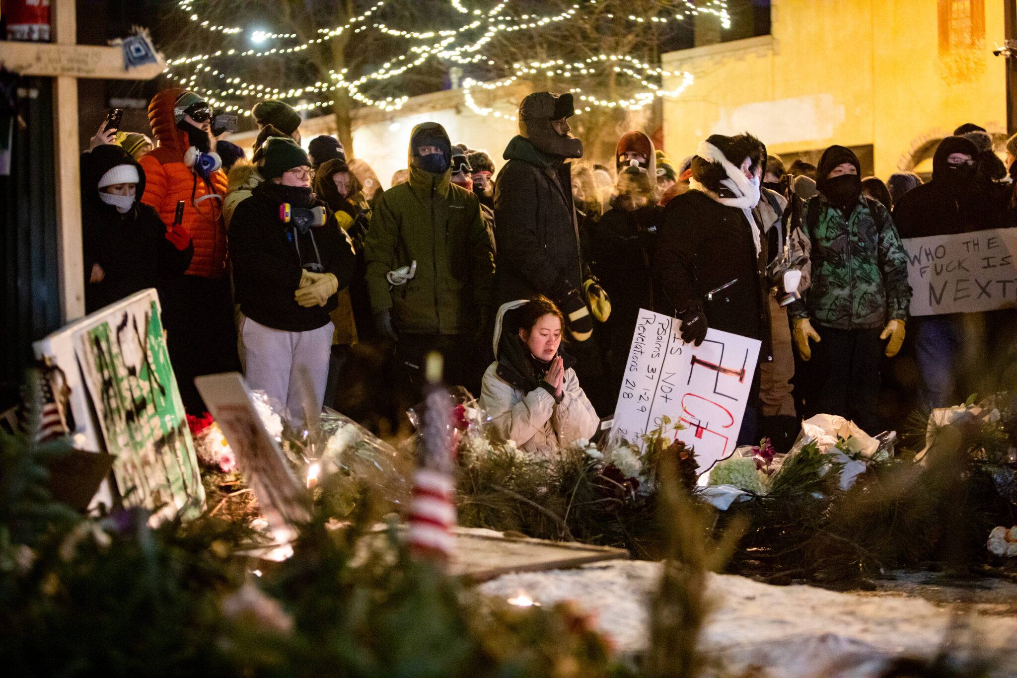 A person kneeling praying, a crowd of people dressed warmly behind them, flowers and candles are in front of them