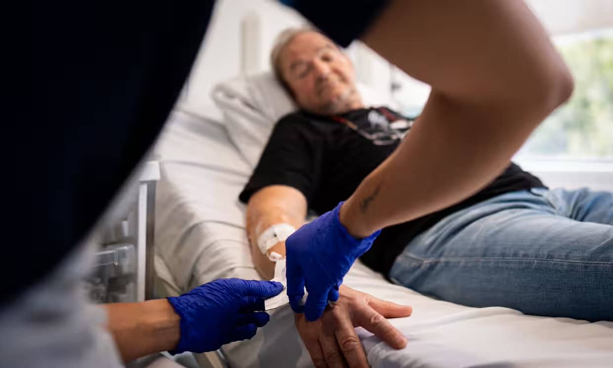 A person wearing jeans and a tshirt laying in a hospital bed as someone wearing blue latex gloves is putting something in an IV coming out of their arm