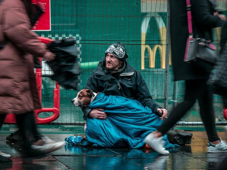 A man sitting with his dog in his lap, the dog is wrapped in a blue blanket as people walk by