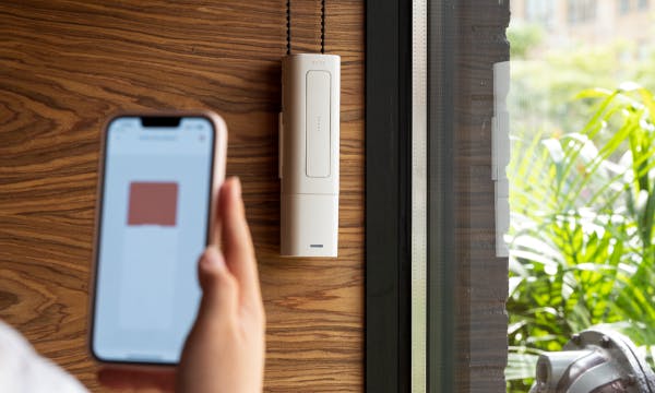 A white remote-sized device attached to a wall next to a window, there's a small black chain going into and out of the device, a person is holding a smartphone in the foreground