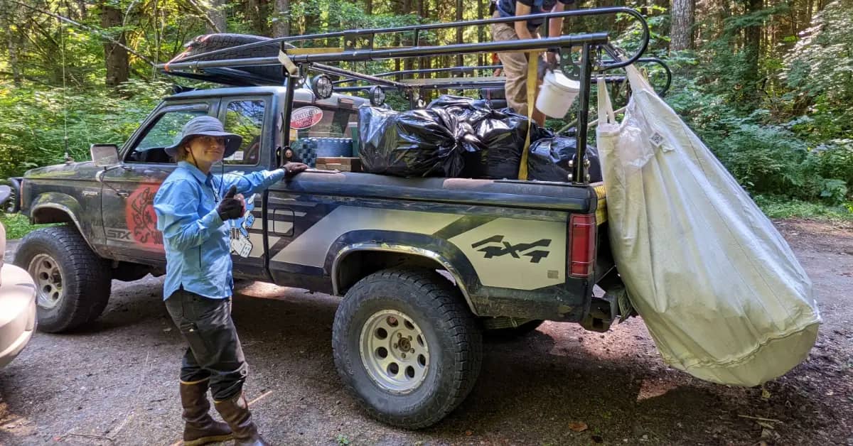 A person wearing boots, pants, a blue shirt, and a bucket hat next to a truck with the bed filled with trash bags