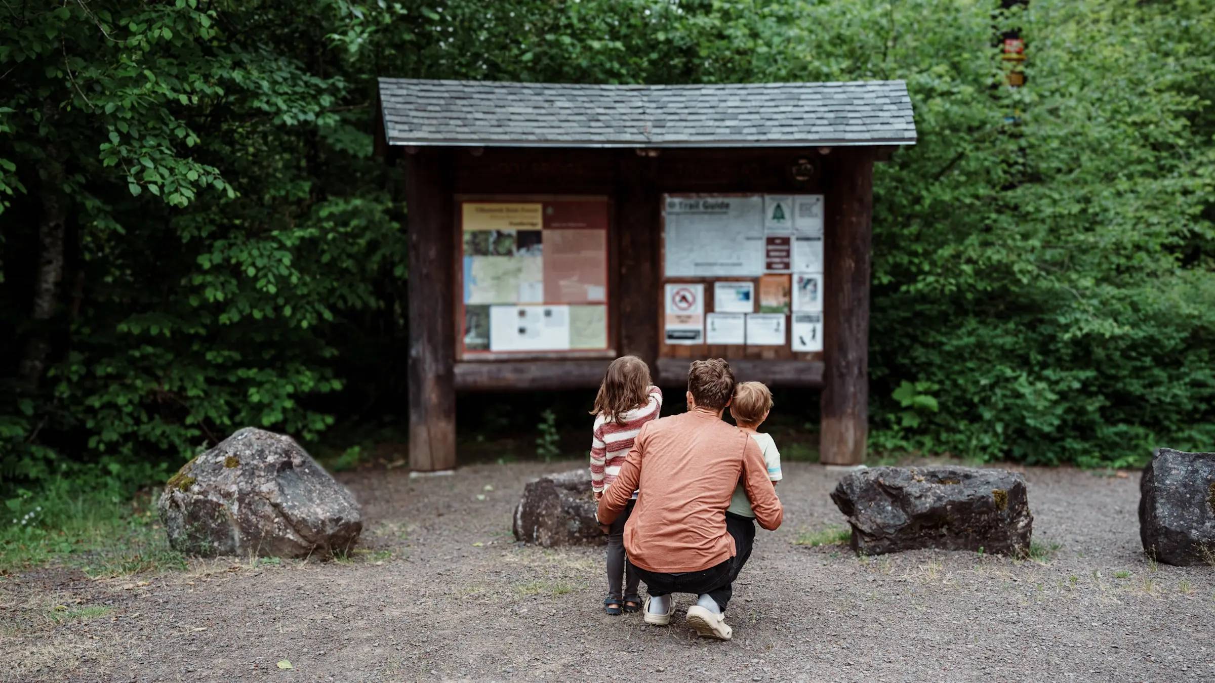 An adult kneeling between two small children, pictured from behind looking at wooden signage in front of a wooded area