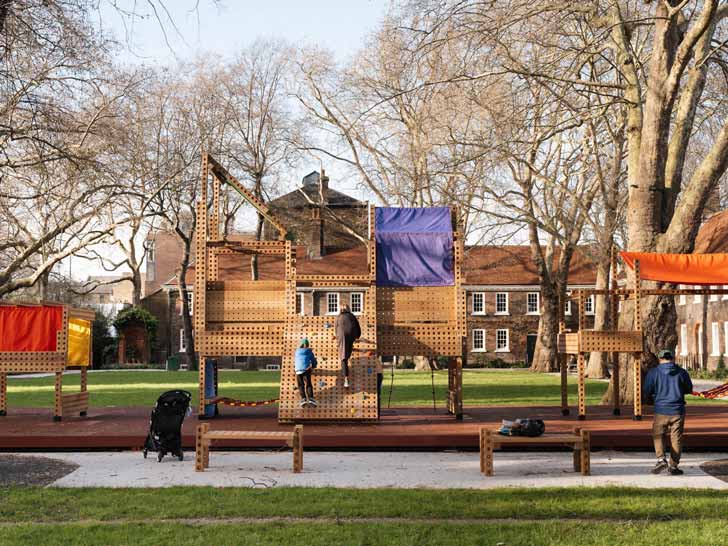 Two kids playing on a wooden play structure, it has holes in it, with colorful climbing stones on the side, and a purple and orange tent covering parts of it
