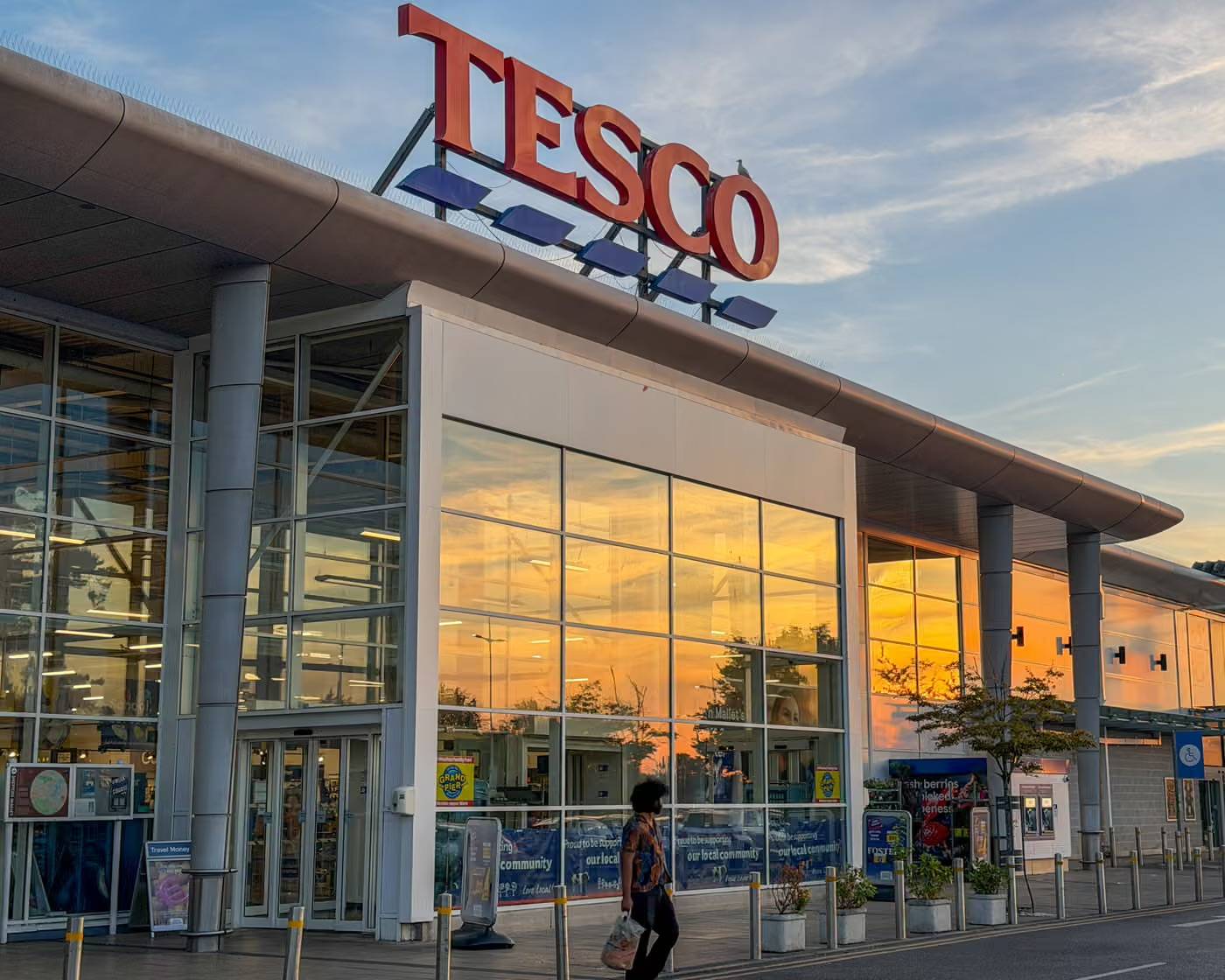 Tesco supermarket storefront at dusk with large illuminated red sign, glass entrance doors, and a person walking past. Golden sunset sky reflects in the windows.