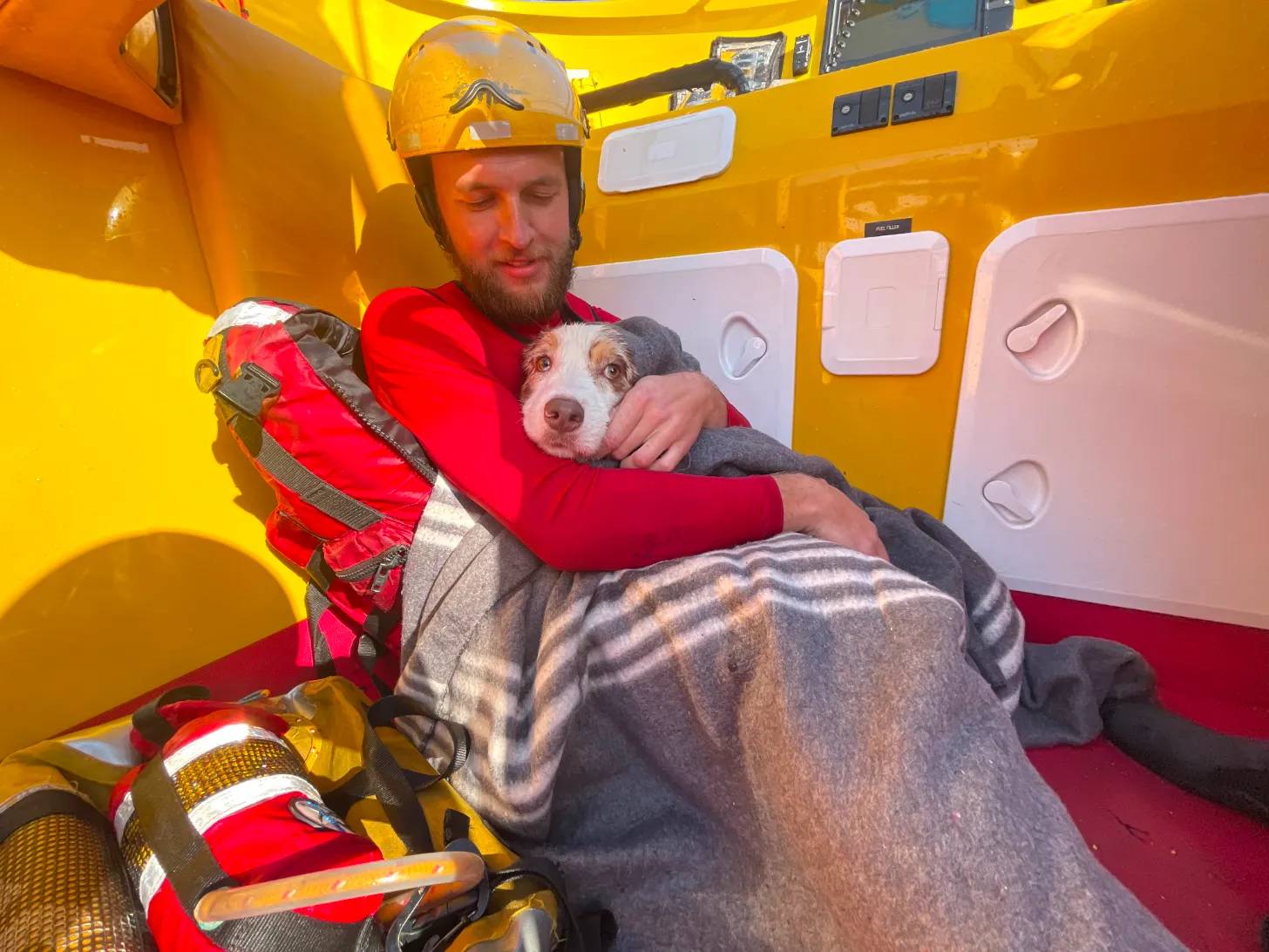 A bearded man wearing a red jacket and yellow safety helmet holds a small brown and white dog wrapped in a gray blanket inside a yellow rescue vehicle with red and white interior panels.