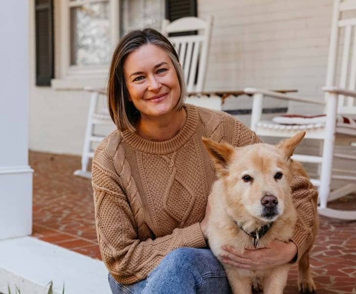 A woman wearing jeans and a tan sweater, sitting with her tan dog on a front porch stoop
