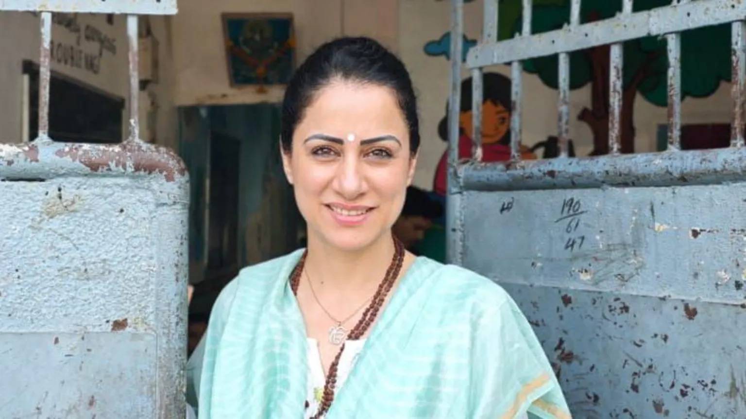 A woman with dark hair pulled back, a teal dress, and brown beaded necklace