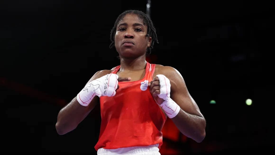 A woman boxer in a red tank top pointing to the refugee team crest on her shirt