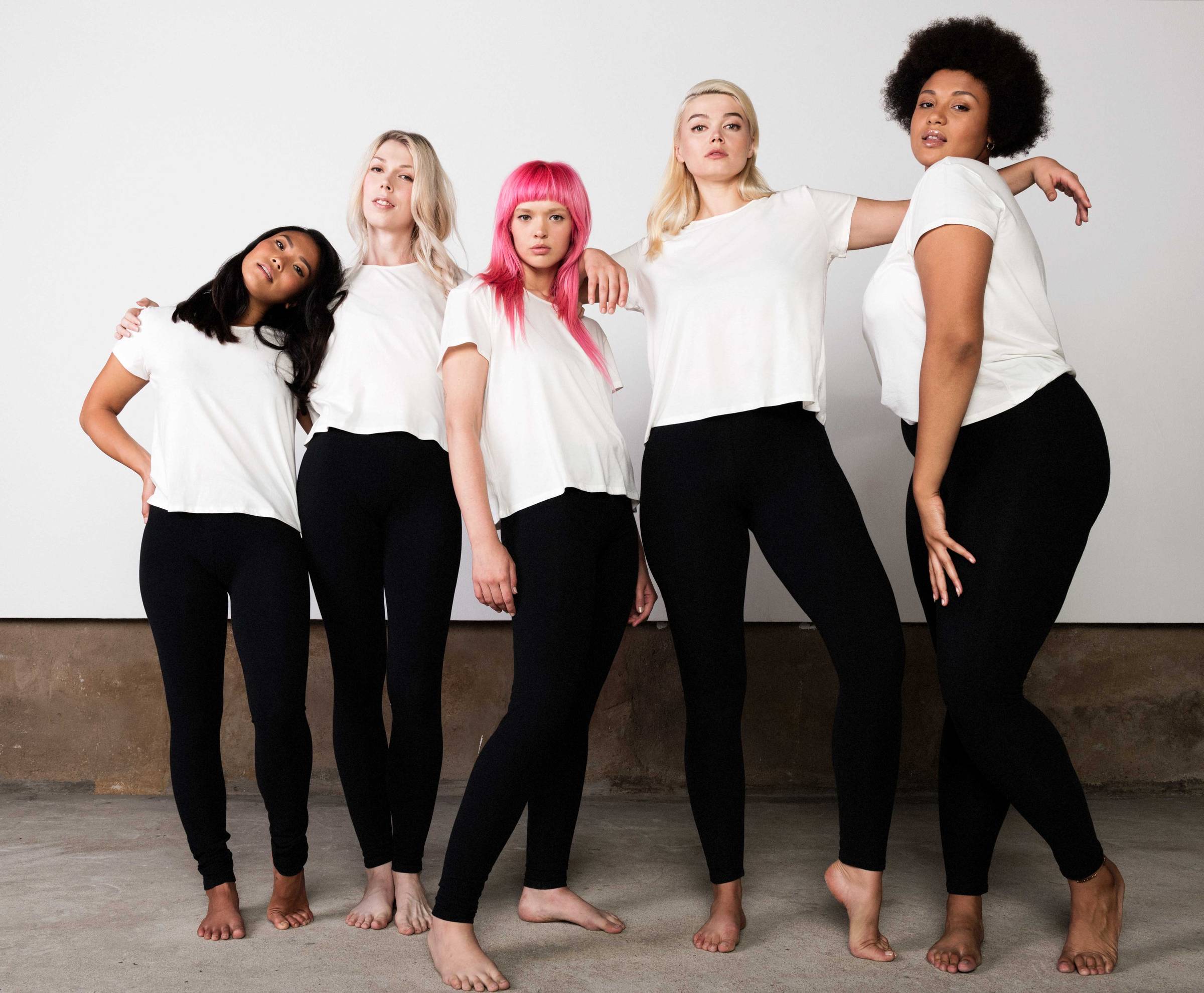 Five people standing together in black leggings and white t-shirts on a concrete floor and a white wall background