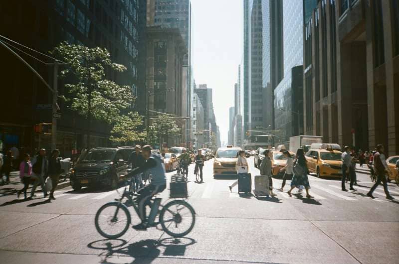 A cyclist rides through a busy urban street lined with tall buildings, yellow taxis, and pedestrians crossing intersections.