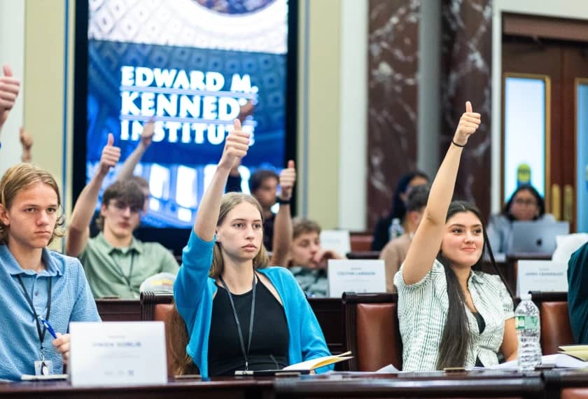 Students seated at a table with name placards raise their hands during a panel or presentation event. A banner reading "EDWARD M. KENNEDY INSTITUTE" is visible on the wall behind them.