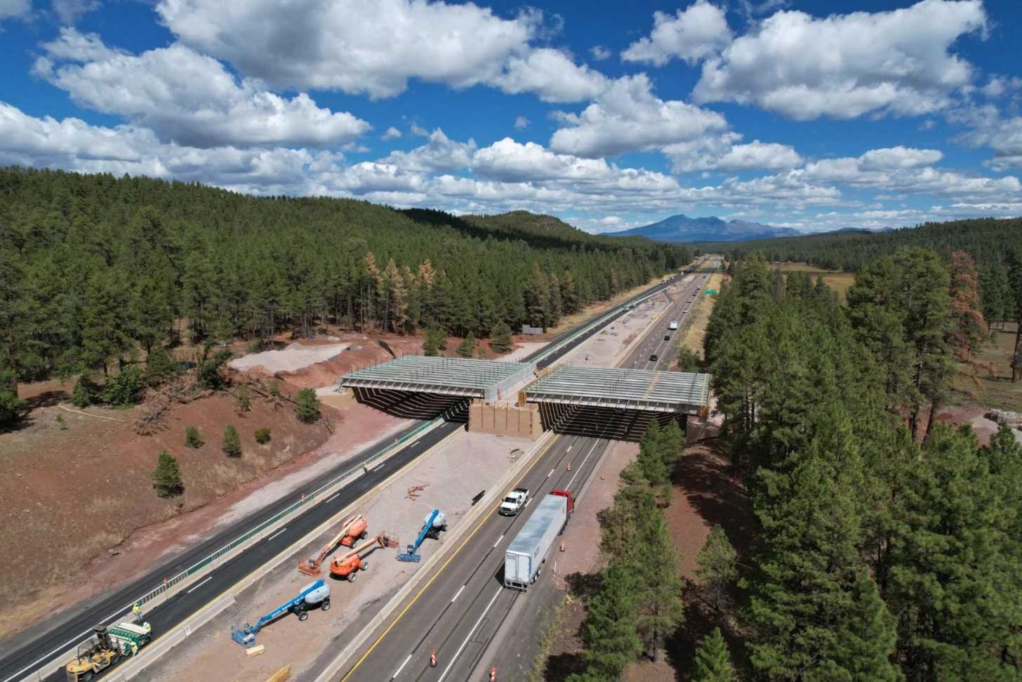 Aerial view of an overpass under construction over a roadway, trees line each side of it