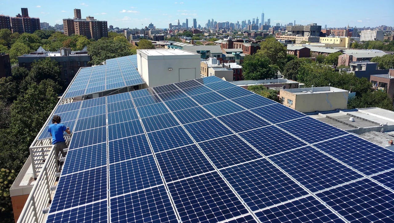 A rooftop filled with solar panels, a person in a blue shirt is working on one of them, a tree canopy and other building roofs surround the building