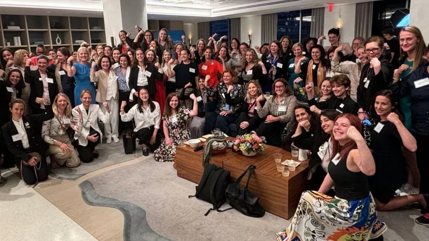 A large group photo of women standing in a large room, most of them are holding one arm up like "Rosie the Riveter"