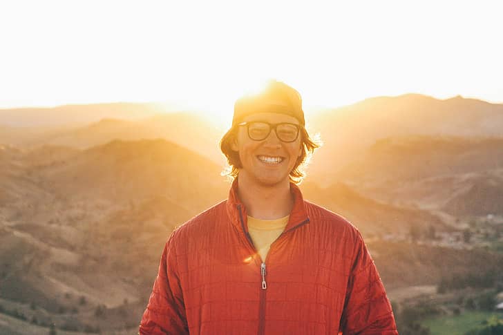 Person in a backwards hat and red jacket standing with the sun and mountains behind them