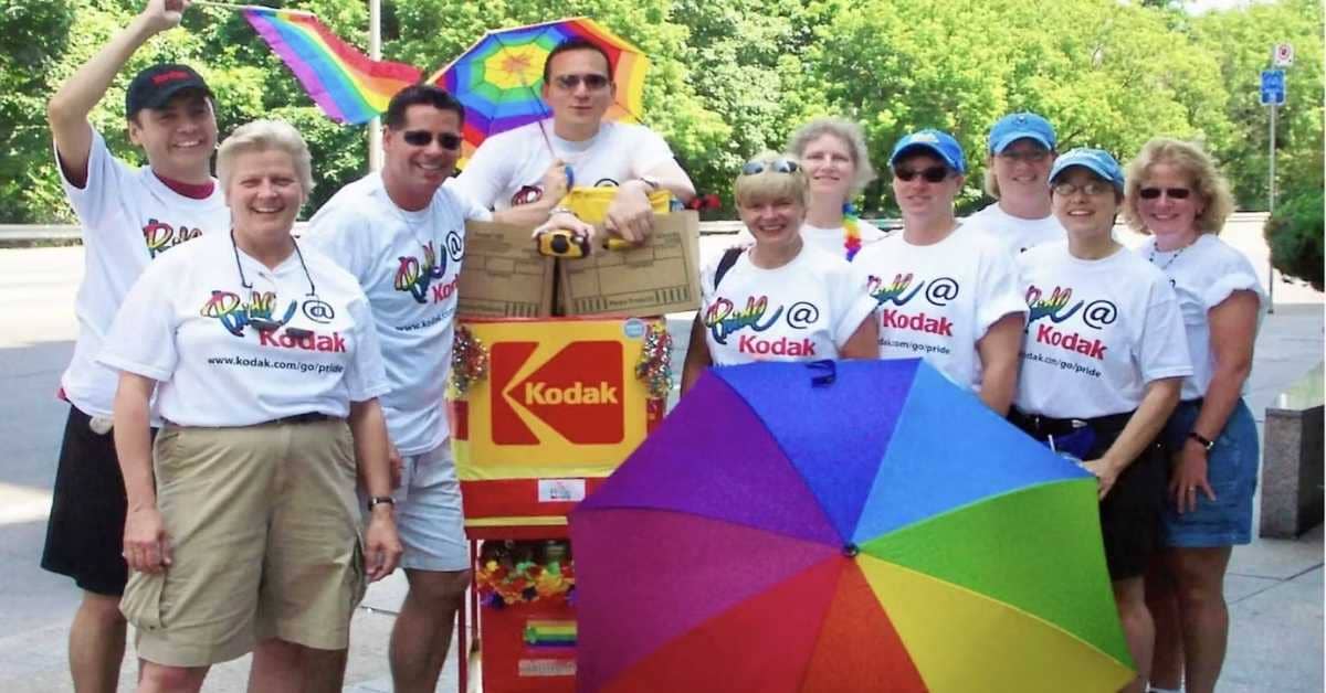 A group of about 10 people wearing "Pride @ Kodak" shirts, rainbow umbrellas, a Kodak box