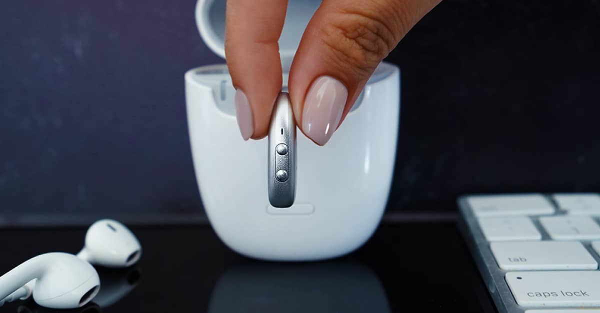 A thumb and forefinger holding a silver hearing aid in front of a white case