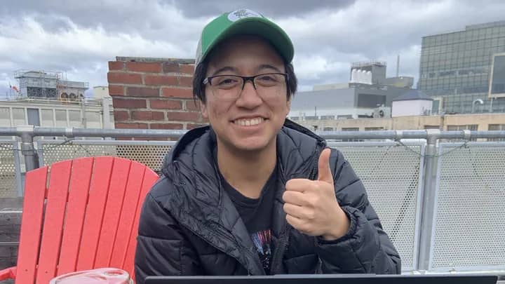 A person wearing a green baseball hat and black jacket smiling and giving a thumbs up