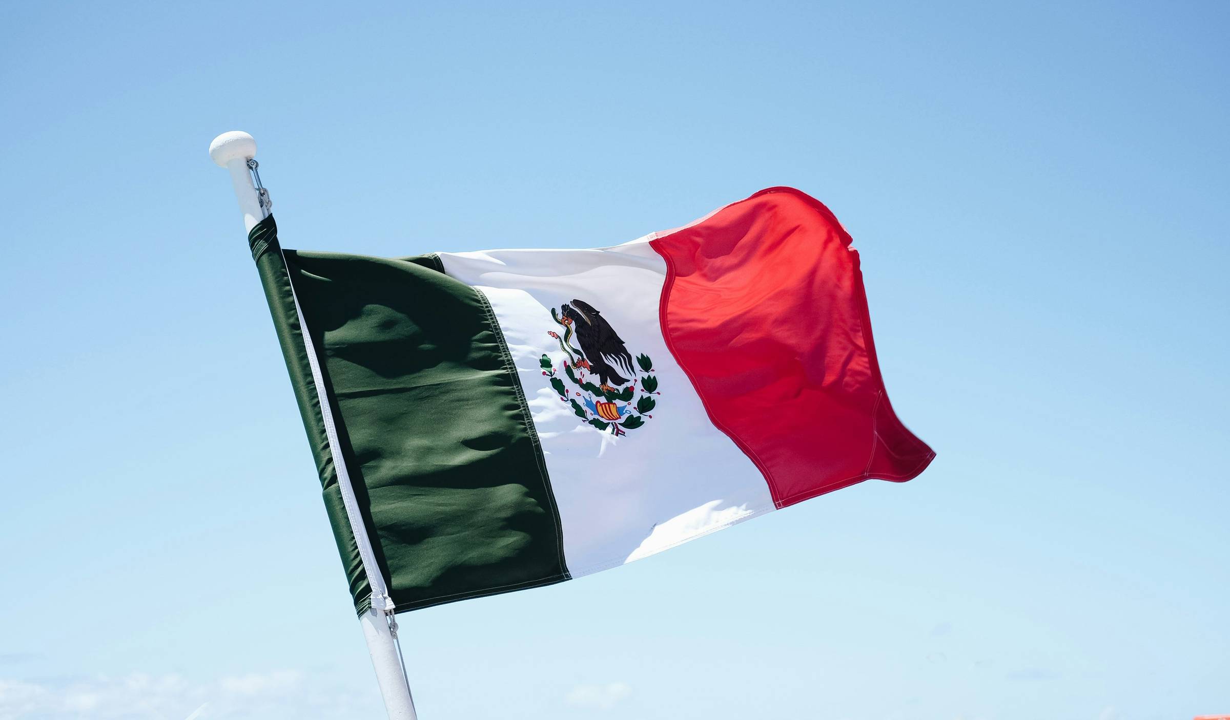 The Mexican flag waves against a clear blue sky, showing three vertical stripes in green, white, and red, with the national eagle emblem in the center of the white stripe.