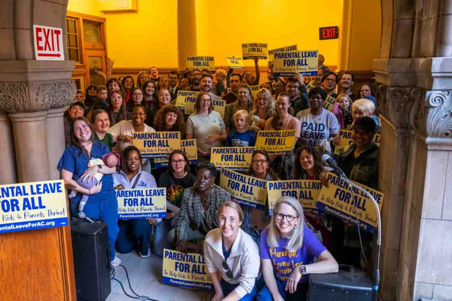 A large group of people holding yellow and blue signs reading "PARENTAL LEAVE FOR ALL" gather indoors beneath yellow walls and an EXIT sign. Several people hold infants. The signs include text reading "To Help Kids & Parents Thrive" and "ParentalLeaveForAC.org".