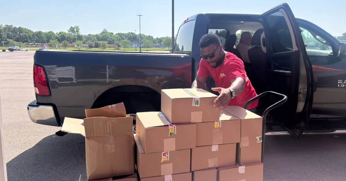 A Black man wearing a red tee and sunglasses unloading cardboard boxes from a black truck