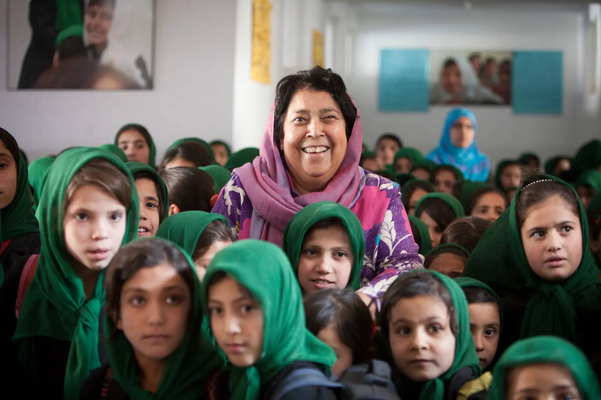 A woman wearing a pink headscarf smiling, she's surrounded by young girls in green headscarfs