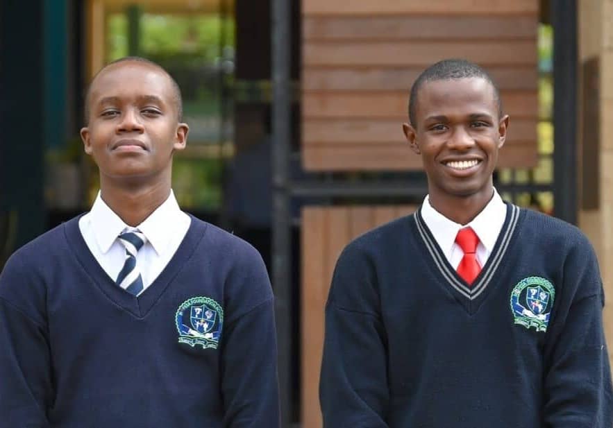 Two people wearing blue sweaters, white shirts, and ties smiling at the camera