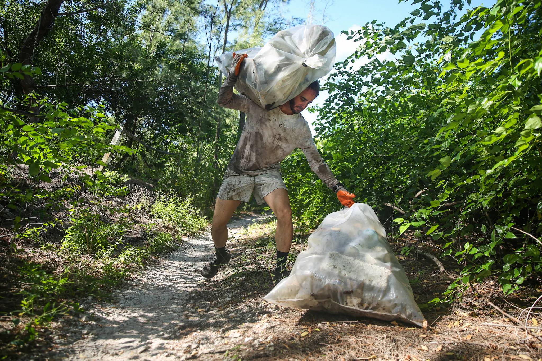 A man in dirty grey shorts and shirt carrying two large trash bags in a forested area