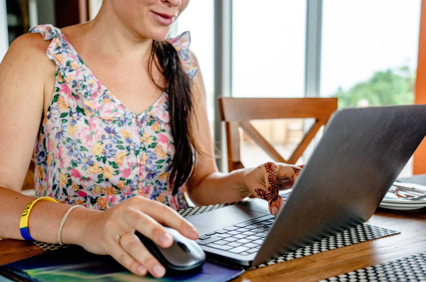 A person wearing a colorful floral tank top uses a laptop and computer mouse at a wooden table indoors, with windows visible in the background.