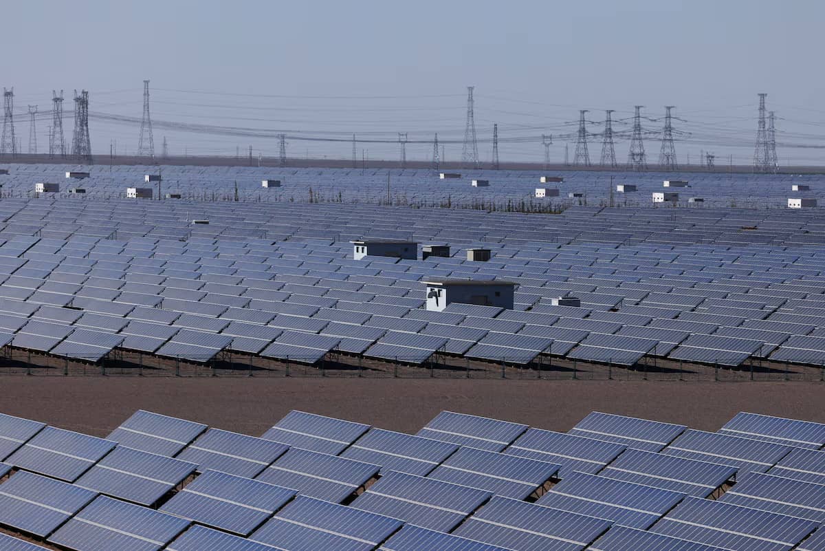 Vast solar panel array stretching across flat terrain with electrical transmission towers visible in the hazy background.
