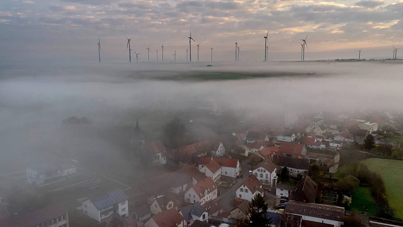 Homes covered in thick fog, a field of wind turbines is visible in the distance