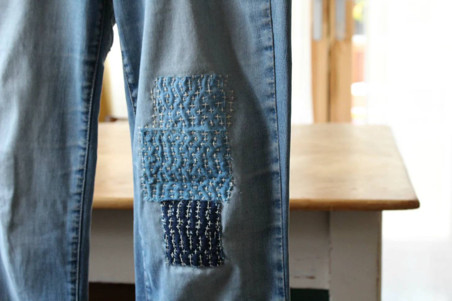 Blue jeans with a patched area on the thigh featuring cross-stitch embroidery in light blue and dark blue thread.