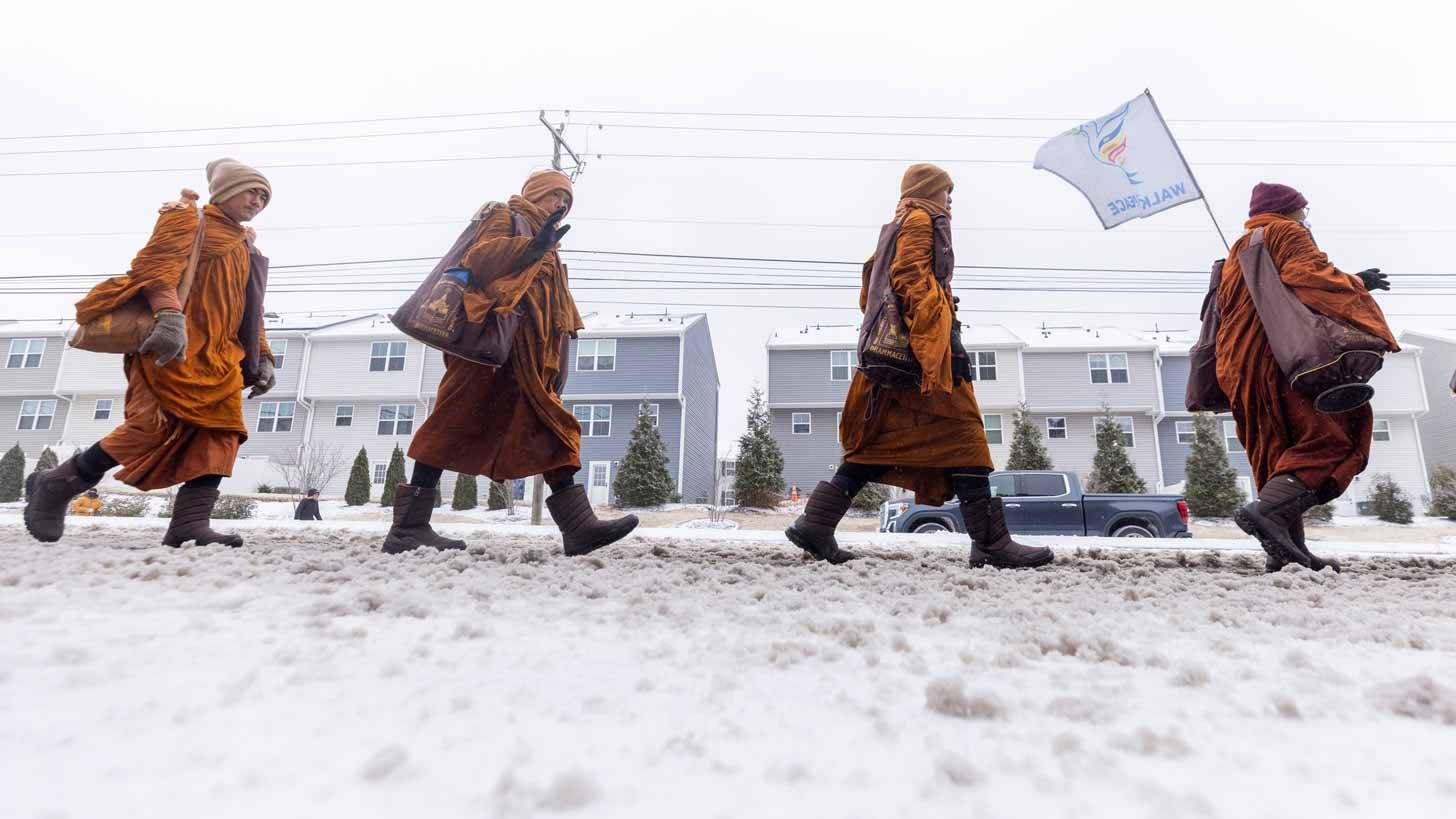 Four monks wearing orange robes, winter hats, and snow boots walking through snow