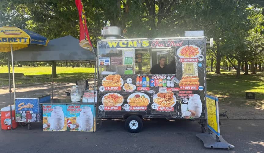 Food truck parked in a tree-lined park with a black canopy tent beside it. The truck displays menu items including empanadas, hot dogs, and other prepared foods with prices visible on its exterior.