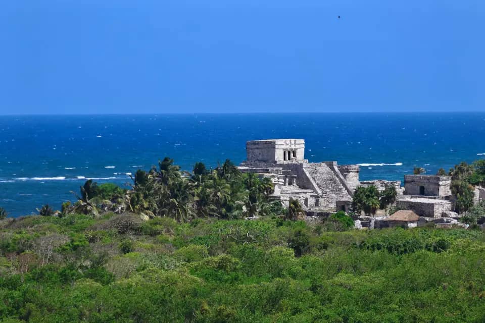 Ruins visible above a green tree canopy with the blue ocean in the background