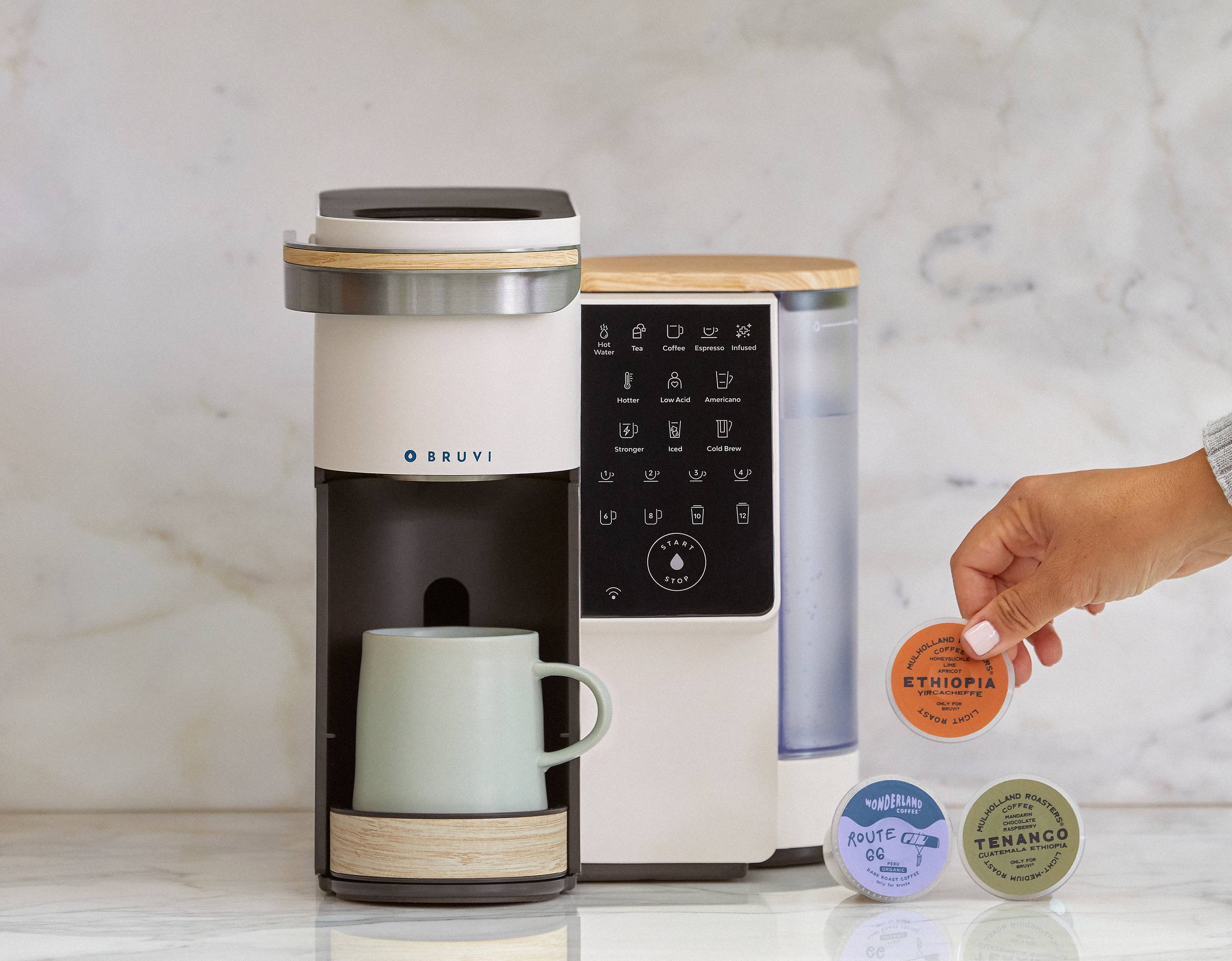 A white, wood, silver, and black single-serve coffee maker on a marble tabletop with a hand holding a pod of coffee next to two others resting on the counter