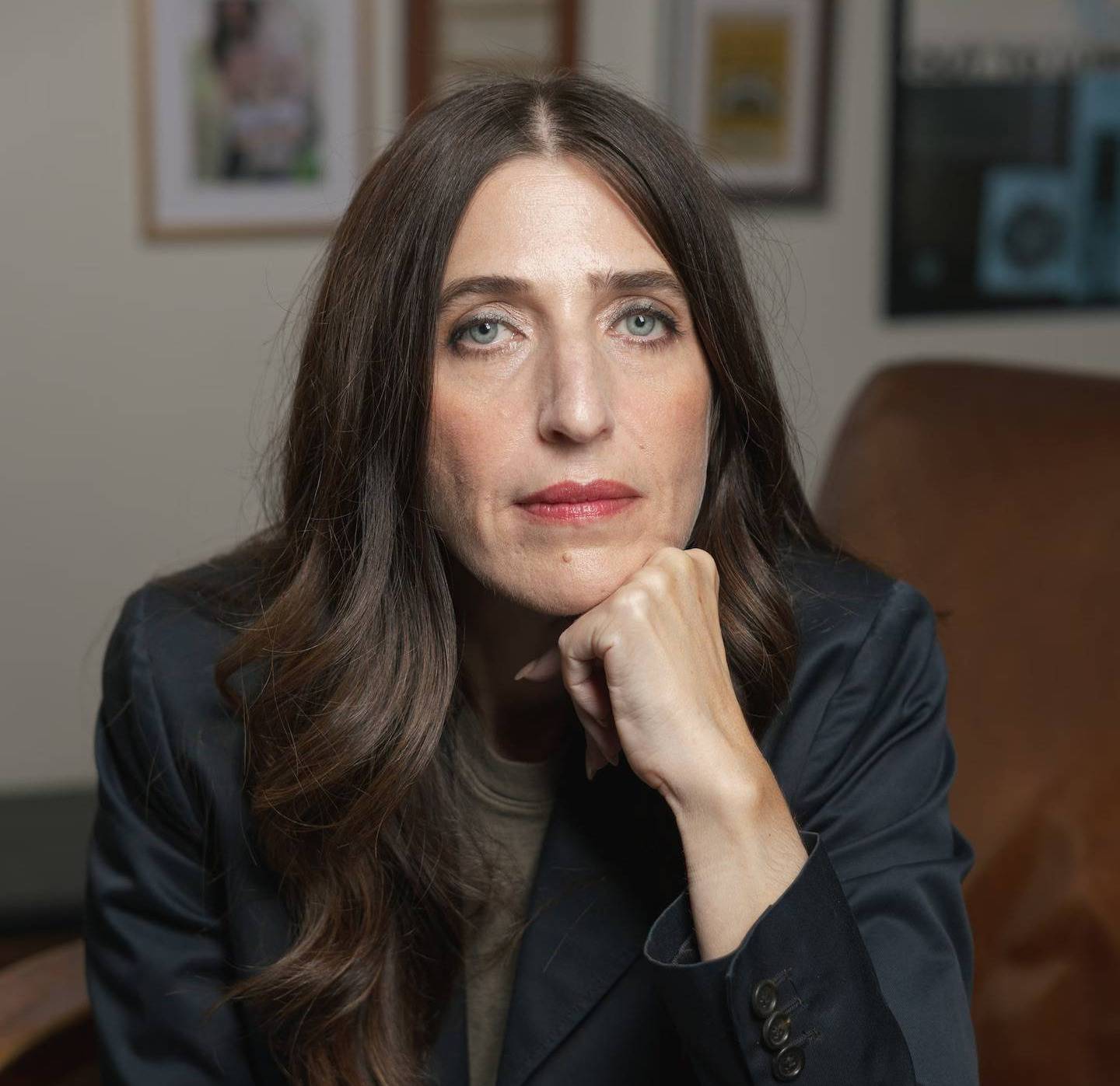Woman with long dark hair wearing a dark blazer, resting her chin on her hand, seated at a wooden table in an office setting with framed pictures visible on the wall behind her.
