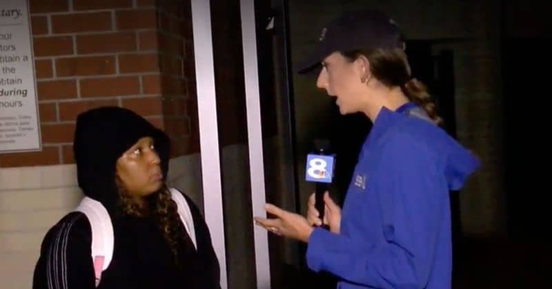 A news person in a blue jacket holding a microphone talking to a person in a black hoodie wearing a backpack