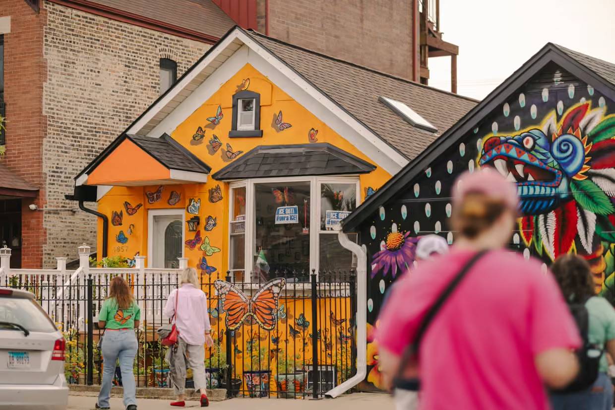 A bright orange house with butterfly murals on its walls and gable, featuring a black roof and white trim. Several people stand on the sidewalk in front viewing the building. A colorful dragon mural is visible on an adjacent structure to the right.
