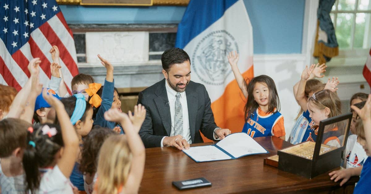 Zohran Mamdani sitting at a desk, kids in Knicks gear are standing around him raising their hands