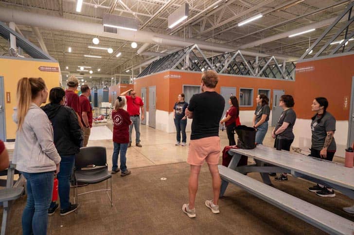 Volunteers gathered listening to someone talk, tiny homes are behind them in a large warehouse